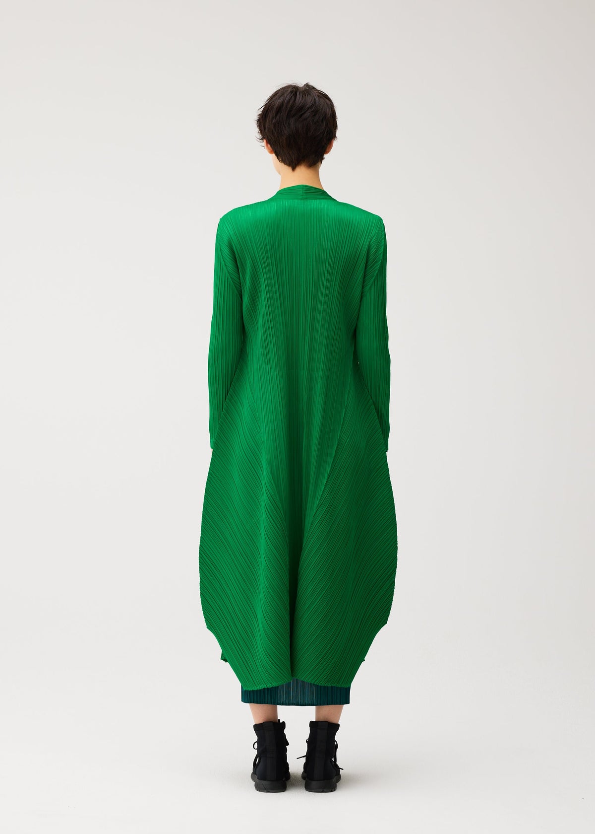 Zoomed view Wearing the MONTHLY COLORS : OCTOBER dress by PLEATS PLEASE ISSEY MIYAKE, a person with short dark hair and black boots stands facing away against a plain white background in a pleated green outfit and color block coat.