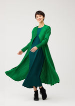 A person wearing the MONTHLY COLORS : OCTOBER coat by PLEATS PLEASE ISSEY MIYAKE over a dark teal pleated dress, smiling mid-step against a white background, paired with black shoes.