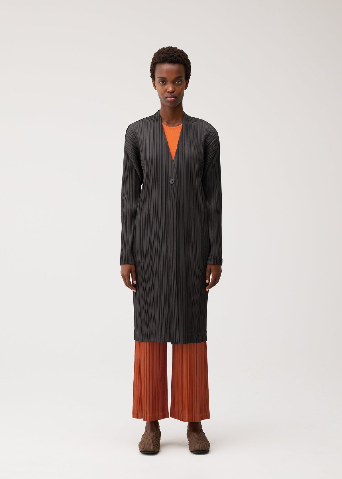 A person stands against a plain light background wearing the PLEATS JACKET by PLEATS PLEASE ISSEY MIYAKE—a black, collarless long jacket crafted from pleated fabric—styled over an orange top and orange pleated pants, with brown shoes. view 4 of 7
