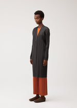 A person stands wearing the PLEATS JACKET by PLEATS PLEASE ISSEY MIYAKE, a long black coat crafted from elegant pleated fabric, layered over an orange shirt and orange pleated pants. They have short curly hair and brown shoes, posed against a plain light background.