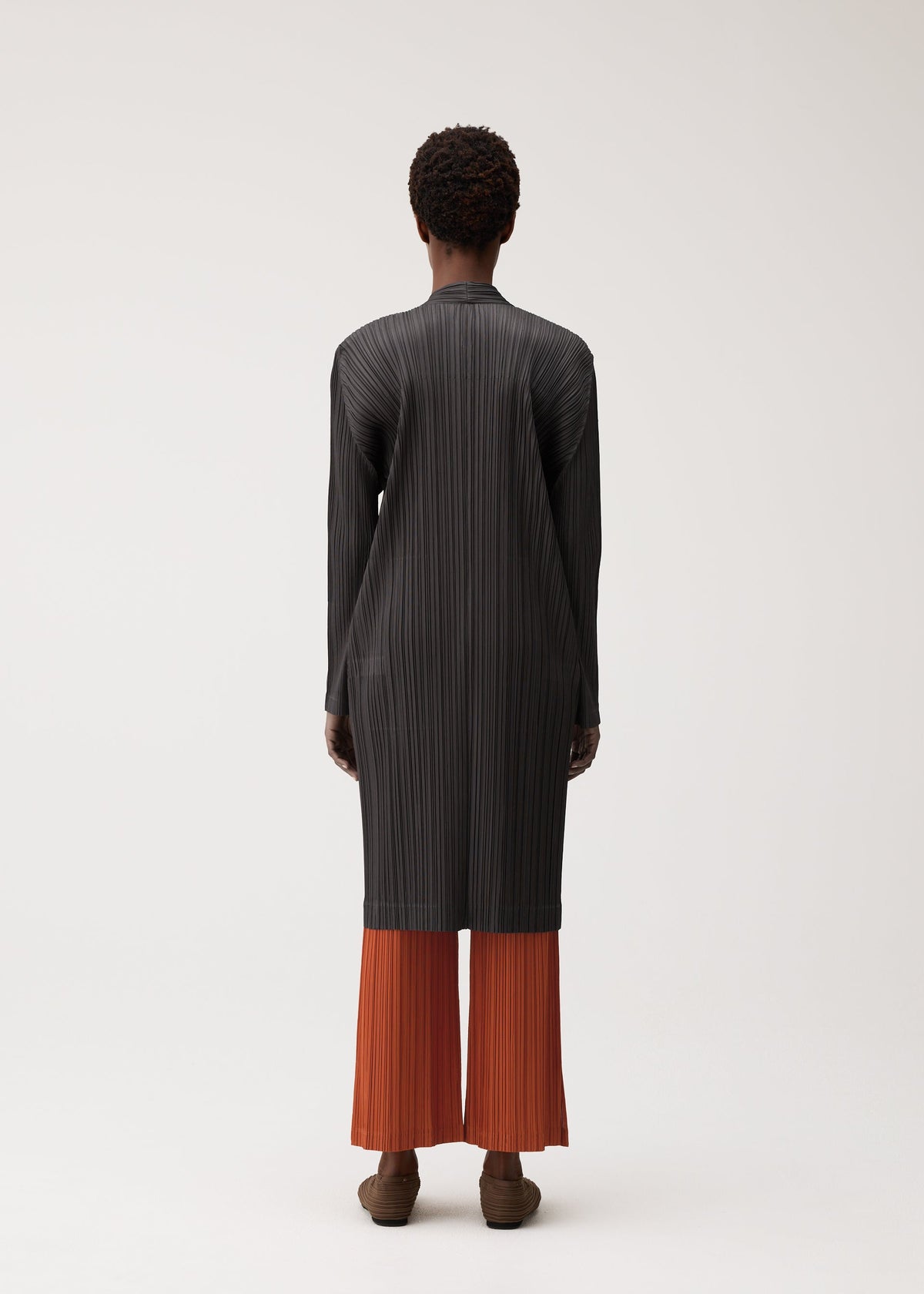 A person stands facing away, dressed in the PLEATS PLEASE ISSEY MIYAKE PLEATS JACKET—a long, black, textured, collarless jacket—layered over loose orange pleated pants with short curly hair and brown shoes, set against a plain light background. view 6 of 7