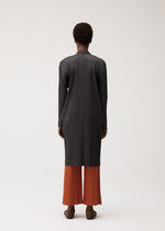 A person stands facing away, dressed in the PLEATS PLEASE ISSEY MIYAKE PLEATS JACKET—a long, black, textured, collarless jacket—layered over loose orange pleated pants with short curly hair and brown shoes, set against a plain light background.