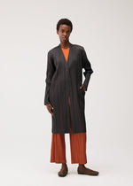 A person stands against a plain background wearing the PLEATS JACKET by PLEATS PLEASE ISSEY MIYAKE, a collarless long jacket crafted from black pleated fabric, layered over an orange pleated outfit and paired with brown shoes; they have one hand in their pocket and display a relaxed posture.