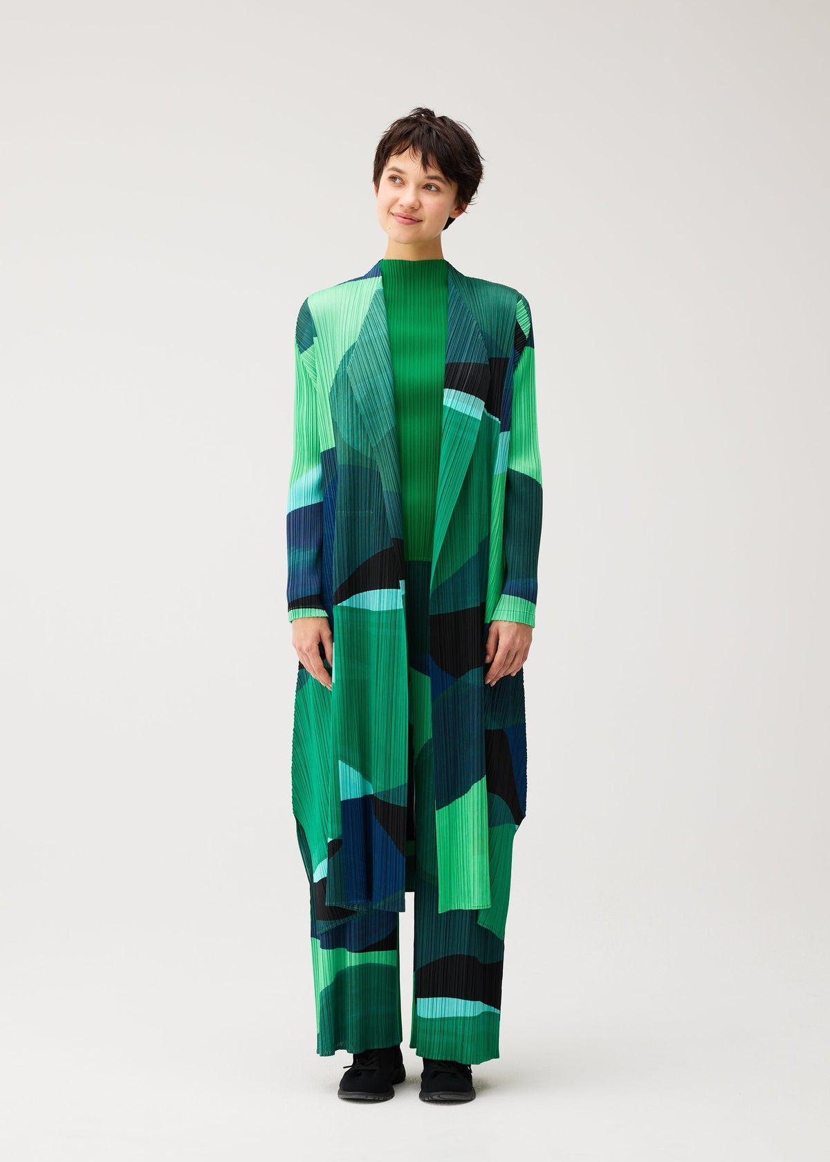 Zoomed view A person stands smiling in PLEATS PLEASE ISSEY MIYAKE ORBIT—a long coat with sheer geometric green, teal, and blue shapes over matching pants—against a plain light background.