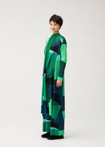 A person with short dark hair wears the ORBIT long geometric-patterned coat by PLEATS PLEASE ISSEY MIYAKE, standing and smiling slightly against a plain light background.