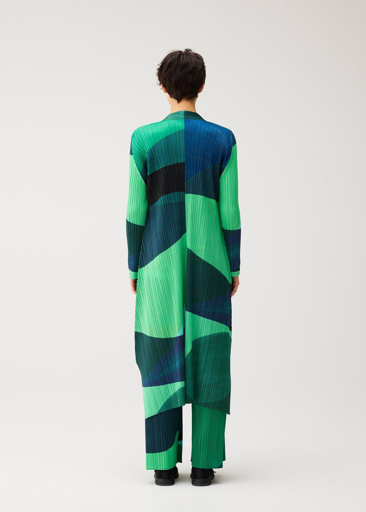 A person stands with their back to the camera, modeling the ORBIT long coat by PLEATS PLEASE ISSEY MIYAKE, featuring sheer colors and an abstract pattern of green, blue, and black shapes on a plain light background. view 7 of 8
