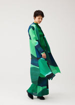 A person in the ORBIT long, pleated green and navy dress with matching wide pants by PLEATS PLEASE ISSEY MIYAKE stands on a white background, looking down with a slight smile.