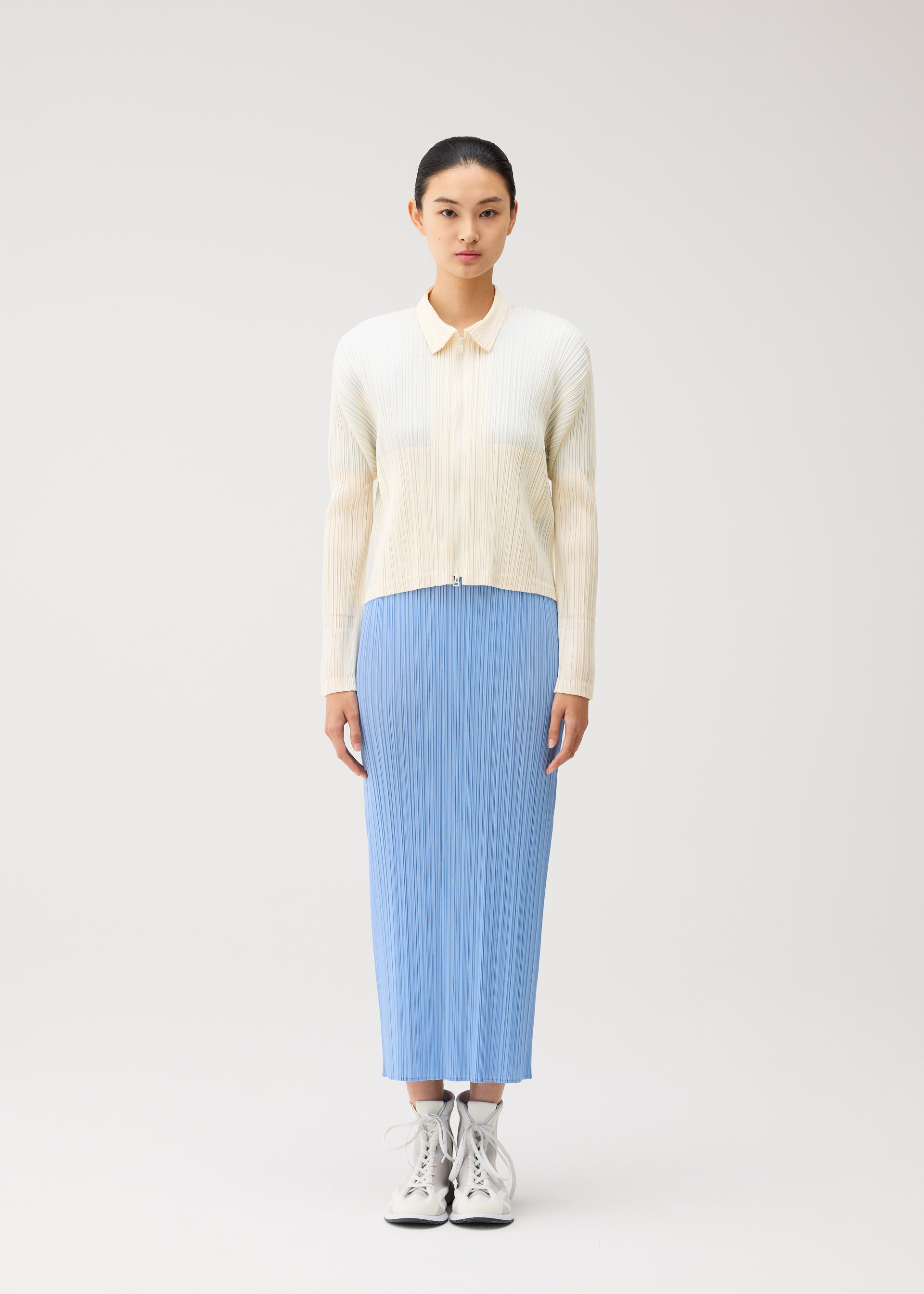 Zoomed view A woman stands against a plain background, wearing the MONTHLY COLORS : AUGUST light blue pleated midi skirt from PLEATS PLEASE ISSEY MIYAKE, styled with a light cream long-sleeve shirt and white high-top sneakers. She faces forward with a neutral expression, capturing the effortless style of midsummer in crisp colors.