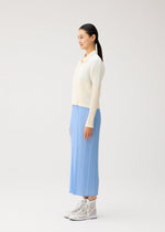 A woman stands in profile against a plain background, wearing the MONTHLY COLORS : AUGUST collection from PLEATS PLEASE ISSEY MIYAKE—a cream zippered cardigan layered over a light blue pleated skirt and paired with gray lace-up boots. Her straight black hair is pulled back in a ponytail, capturing the fresh, vibrant palette of midsummer.