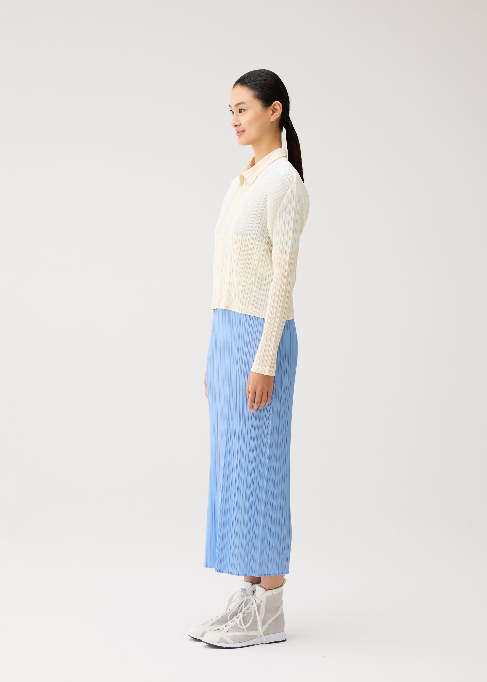 Zoomed view A woman stands in profile against a plain background, wearing the MONTHLY COLORS : AUGUST collection from PLEATS PLEASE ISSEY MIYAKE—a cream zippered cardigan layered over a light blue pleated skirt and paired with gray lace-up boots. Her straight black hair is pulled back in a ponytail, capturing the fresh, vibrant palette of midsummer.