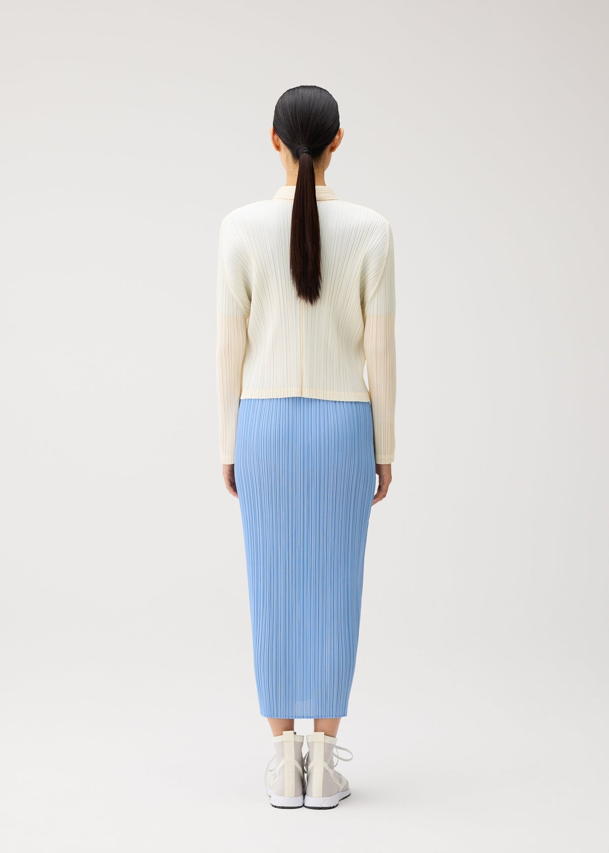 Wearing pieces from PLEATS PLEASE ISSEY MIYAKE’s MONTHLY COLORS: AUGUST collection, a person stands facing away in a cream ribbed top and light blue pleated skirt, paired with white sneakers. Their straight black hair is tied back in a low ponytail. The crisp color palette stands out against the plain, light background for a fresh midsummer effect. view 10 of 11