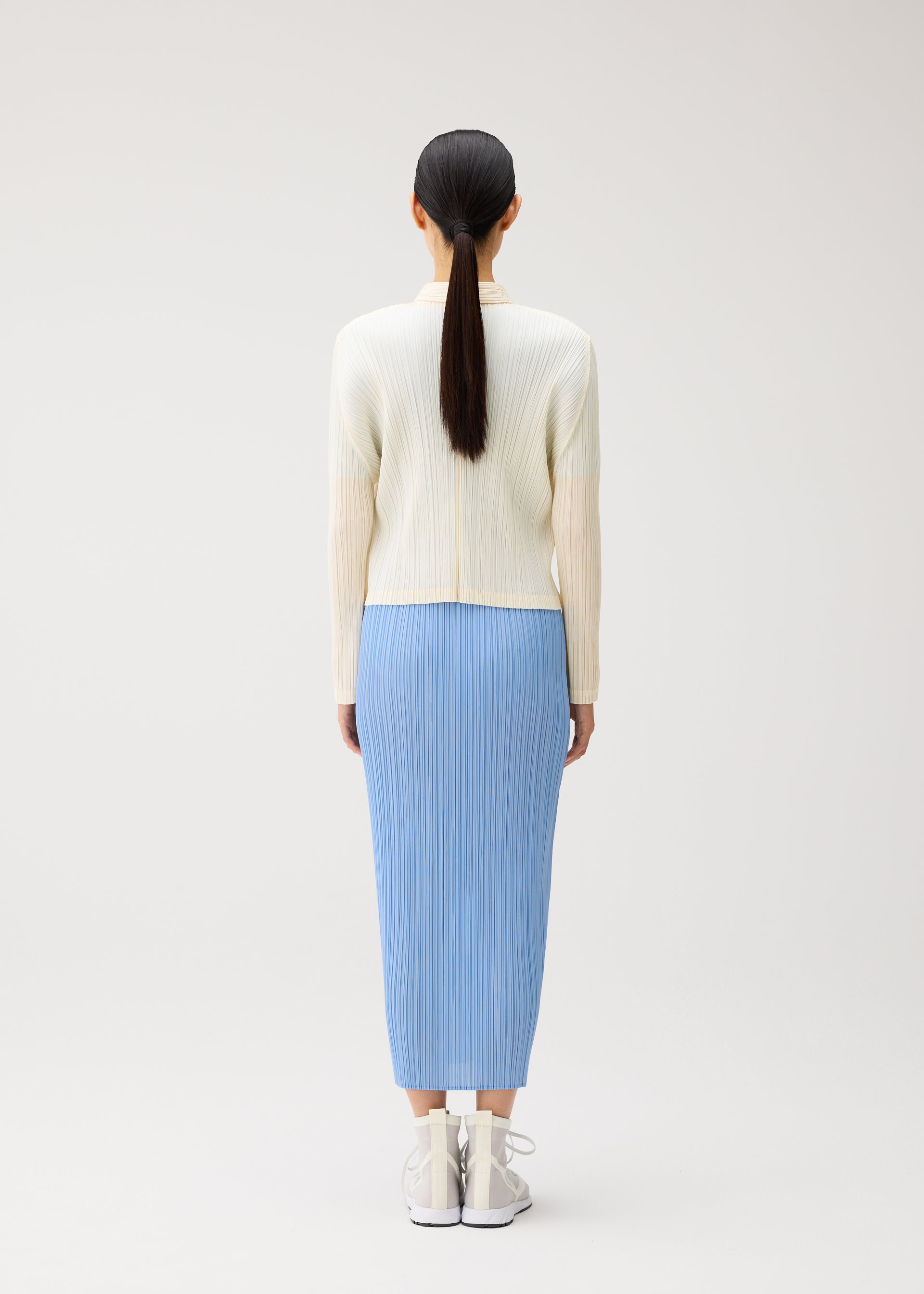 Zoomed view Wearing pieces from PLEATS PLEASE ISSEY MIYAKE’s MONTHLY COLORS: AUGUST collection, a person stands facing away in a cream ribbed top and light blue pleated skirt, paired with white sneakers. Their straight black hair is tied back in a low ponytail. The crisp color palette stands out against the plain, light background for a fresh midsummer effect.