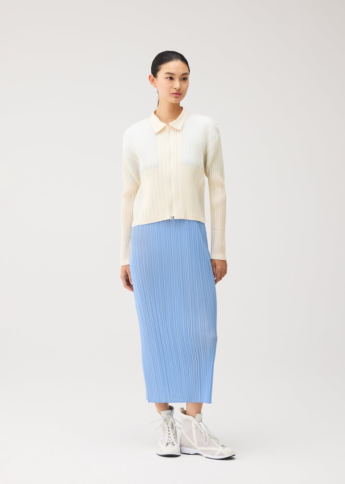 A person stands against a plain background wearing the MONTHLY COLORS : AUGUST cream-colored zippered cardigan from PLEATS PLEASE ISSEY MIYAKE, layered over a light blue pleated skirt, and paired with white high-top sneakers. view 11 of 11