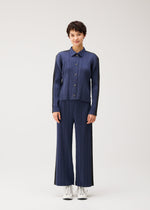 Person stands against a plain background wearing PLEATS PLEASE ISSEY MIYAKE COTTON DENIM—a matching ribbed navy blue button-up shirt and wide-leg pants—paired with white sneakers.