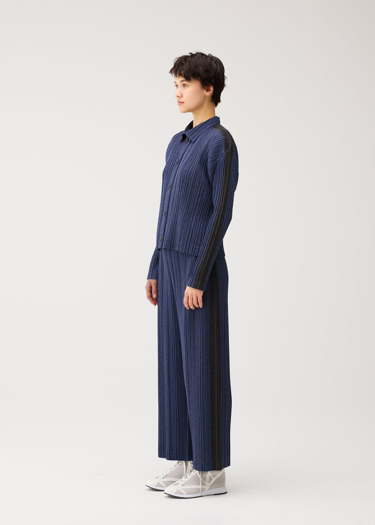 A person stands in profile against a light background, wearing PLEATS PLEASE ISSEY MIYAKE’s COTTON DENIM set—long-sleeved pleated shirt and wide-leg pants—with white high-top sneakers. view 6 of 8