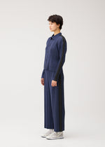 A person stands in profile against a light background, wearing PLEATS PLEASE ISSEY MIYAKE’s COTTON DENIM set—long-sleeved pleated shirt and wide-leg pants—with white high-top sneakers.
