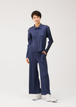 A person wears the PLEATS PLEASE ISSEY MIYAKE COTTON DENIM—a blue, ribbed, button-up shirt with matching wide-leg pants—paired with white sneakers, posing against a plain light background.