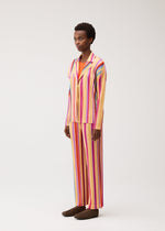 A person stands in a studio against a plain light background, wearing the PLEATS PLEASE ISSEY MIYAKE TAPE TAPE—a coordinated two-piece outfit featuring vertical stripes in pink, orange, yellow, and white—paired with dark brown shoes.