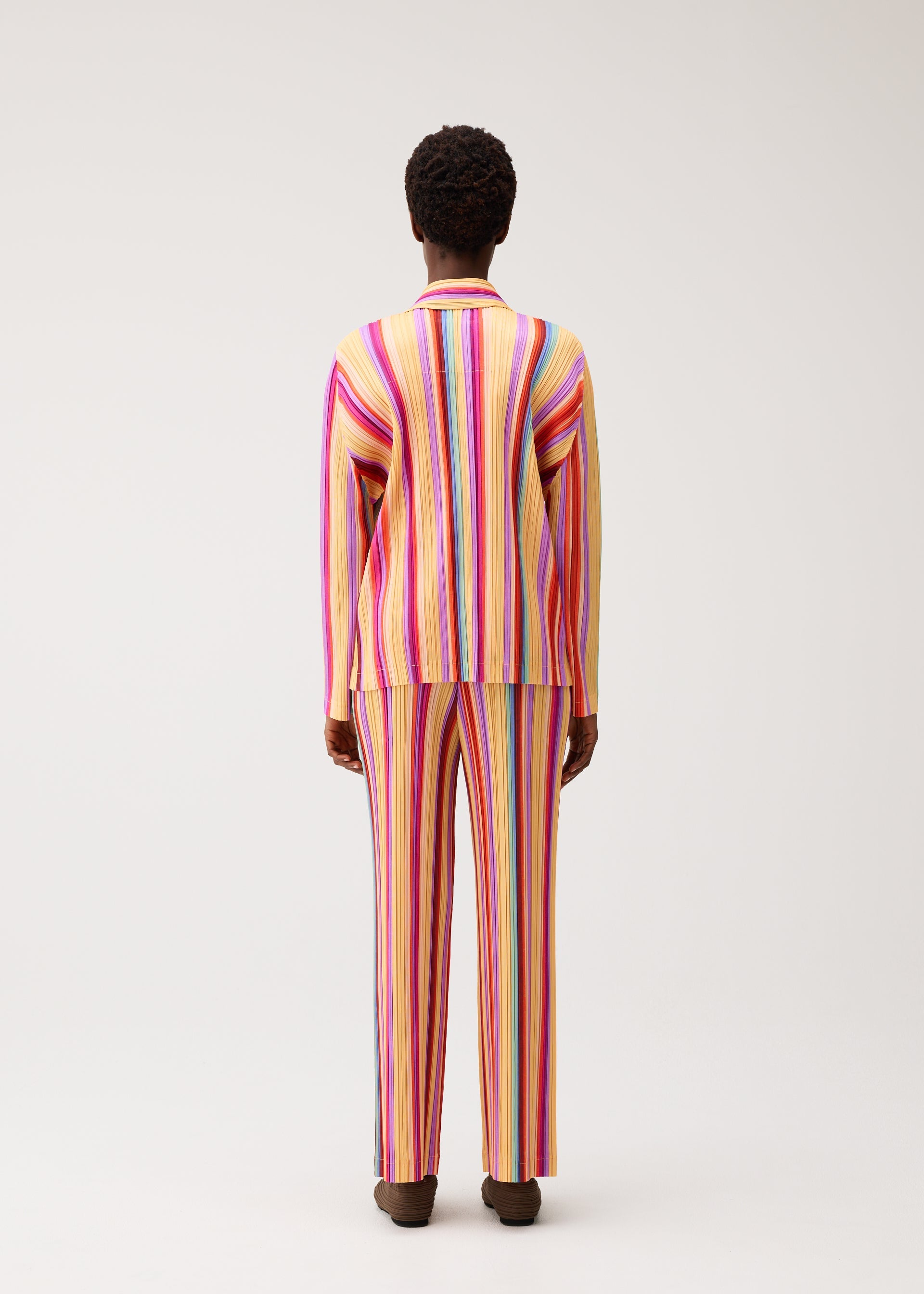 Zoomed view A person stands facing away, wearing the TAPE TAPE two-piece set by PLEATS PLEASE ISSEY MIYAKE—a vibrant ensemble with a matching long-sleeve top and pants featuring vertical stripes in bright hues of pink, yellow, orange, and purple—set against a plain light background.