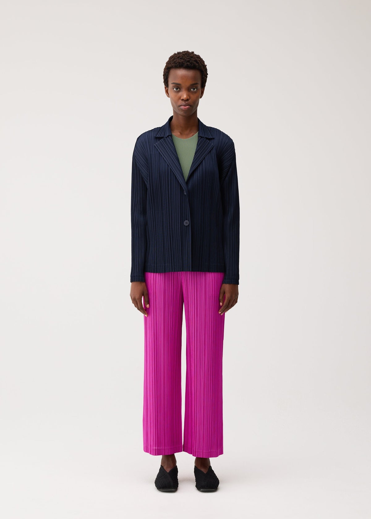A person stands facing forward against a plain light background, wearing the PLEATS PLEASE ISSEY MIYAKE PLEATS JACKET in navy over a green top, paired with bright pink soft pleated pants and black shoes. view 4 of 7