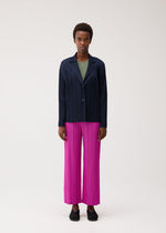 A person stands facing forward against a plain light background, wearing the PLEATS PLEASE ISSEY MIYAKE PLEATS JACKET in navy over a green top, paired with bright pink soft pleated pants and black shoes.