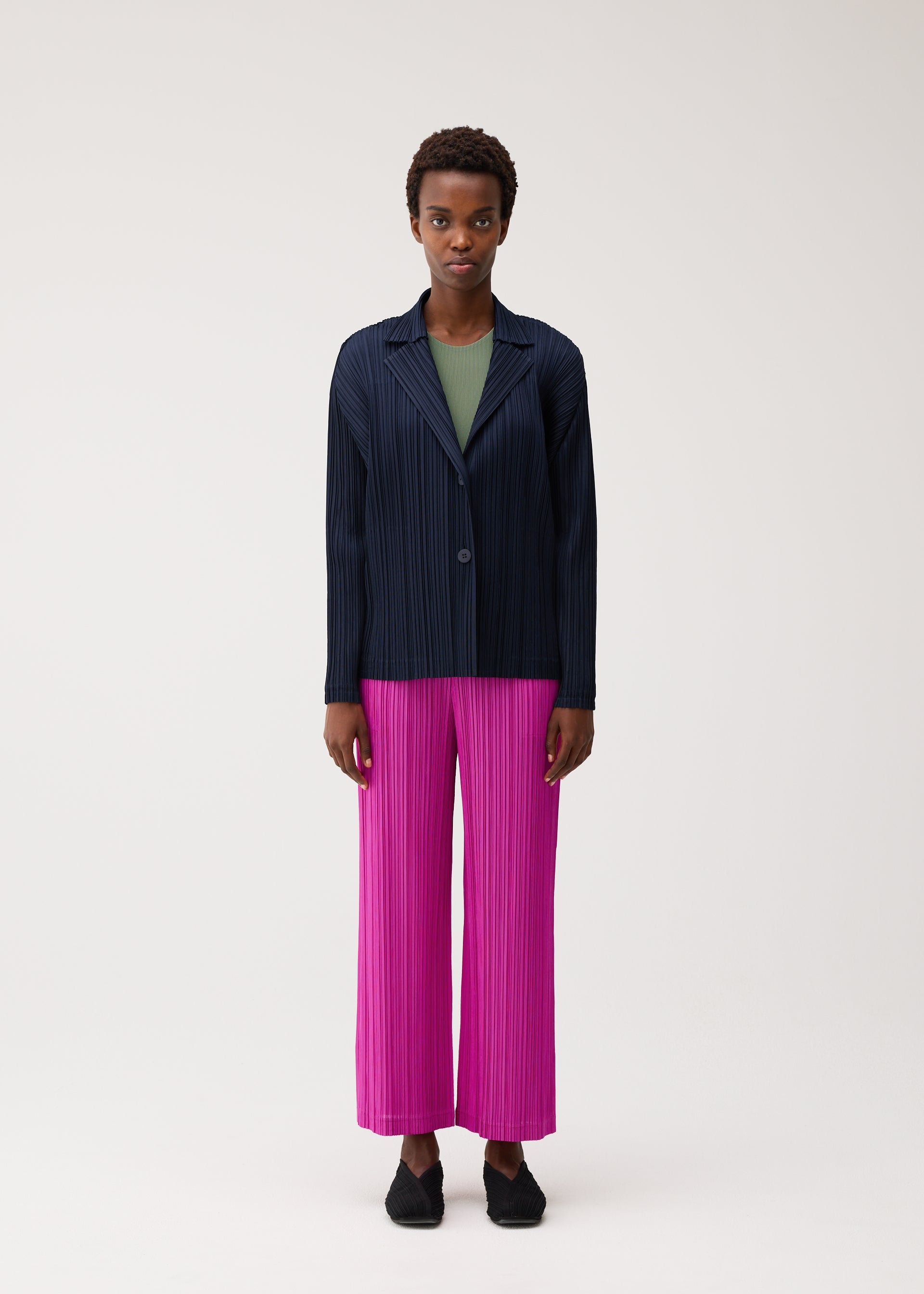Zoomed view A person stands facing forward against a plain light background, wearing the PLEATS PLEASE ISSEY MIYAKE PLEATS JACKET in navy over a green top, paired with bright pink soft pleated pants and black shoes.