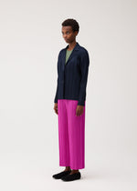 A person stands against a light background wearing the PLEATS JACKET by PLEATS PLEASE ISSEY MIYAKE—a navy blue collared jacket with pockets—paired with a green top, bright pink pleated wide-leg pants, and black shoes.