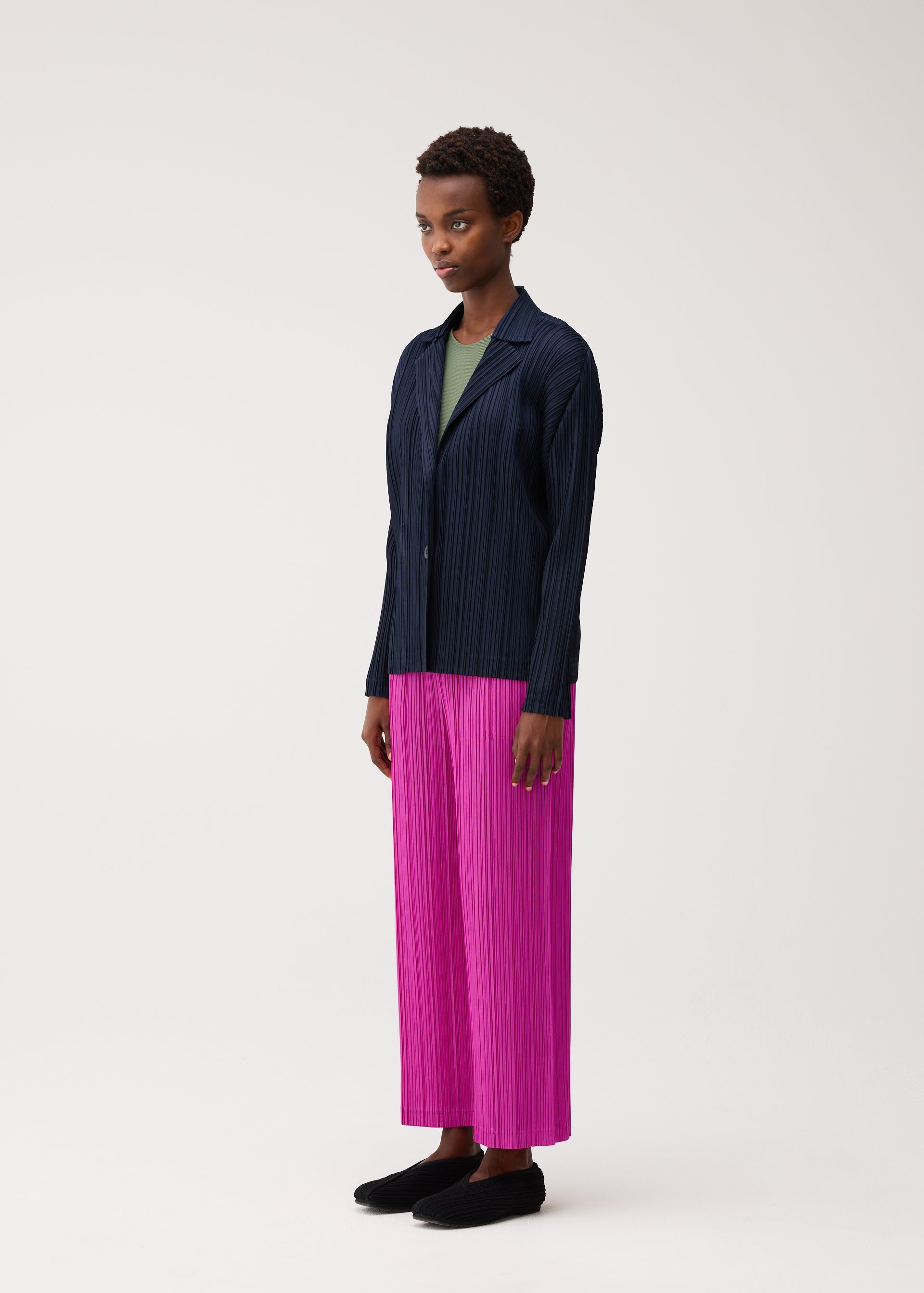 Zoomed view A person stands against a light background wearing the PLEATS JACKET by PLEATS PLEASE ISSEY MIYAKE—a navy blue collared jacket with pockets—paired with a green top, bright pink pleated wide-leg pants, and black shoes.