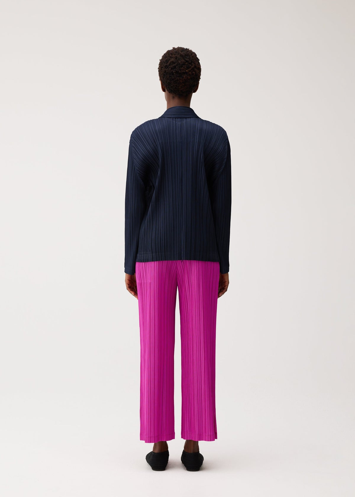 A person with short curly hair is standing facing away on a plain light background, wearing the PLEATS PLEASE ISSEY MIYAKE PLEATS JACKET in navy paired with vibrant pink pleated wide-leg pants and black shoes. view 6 of 7