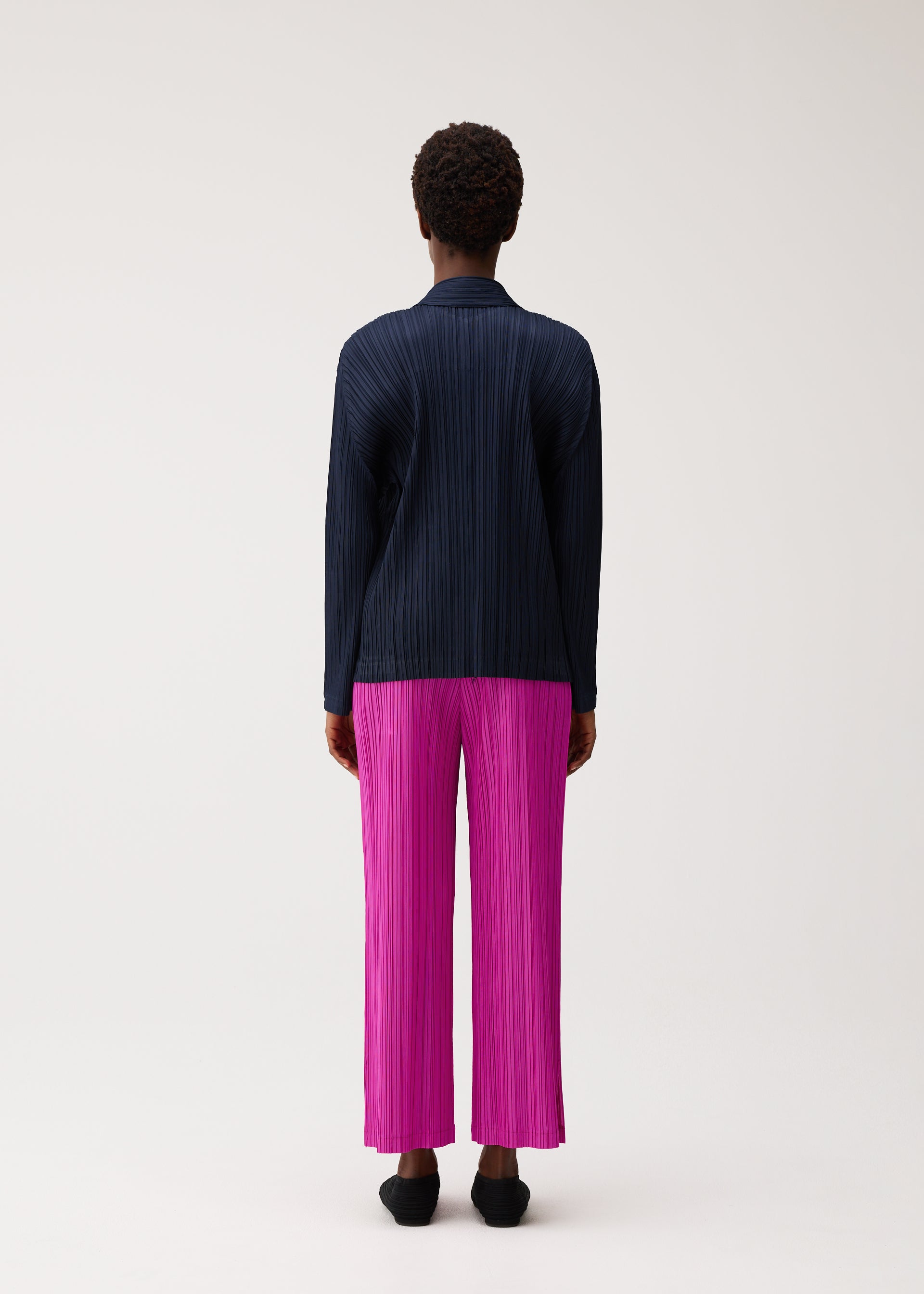 Zoomed view A person with short curly hair is standing facing away on a plain light background, wearing the PLEATS PLEASE ISSEY MIYAKE PLEATS JACKET in navy paired with vibrant pink pleated wide-leg pants and black shoes.