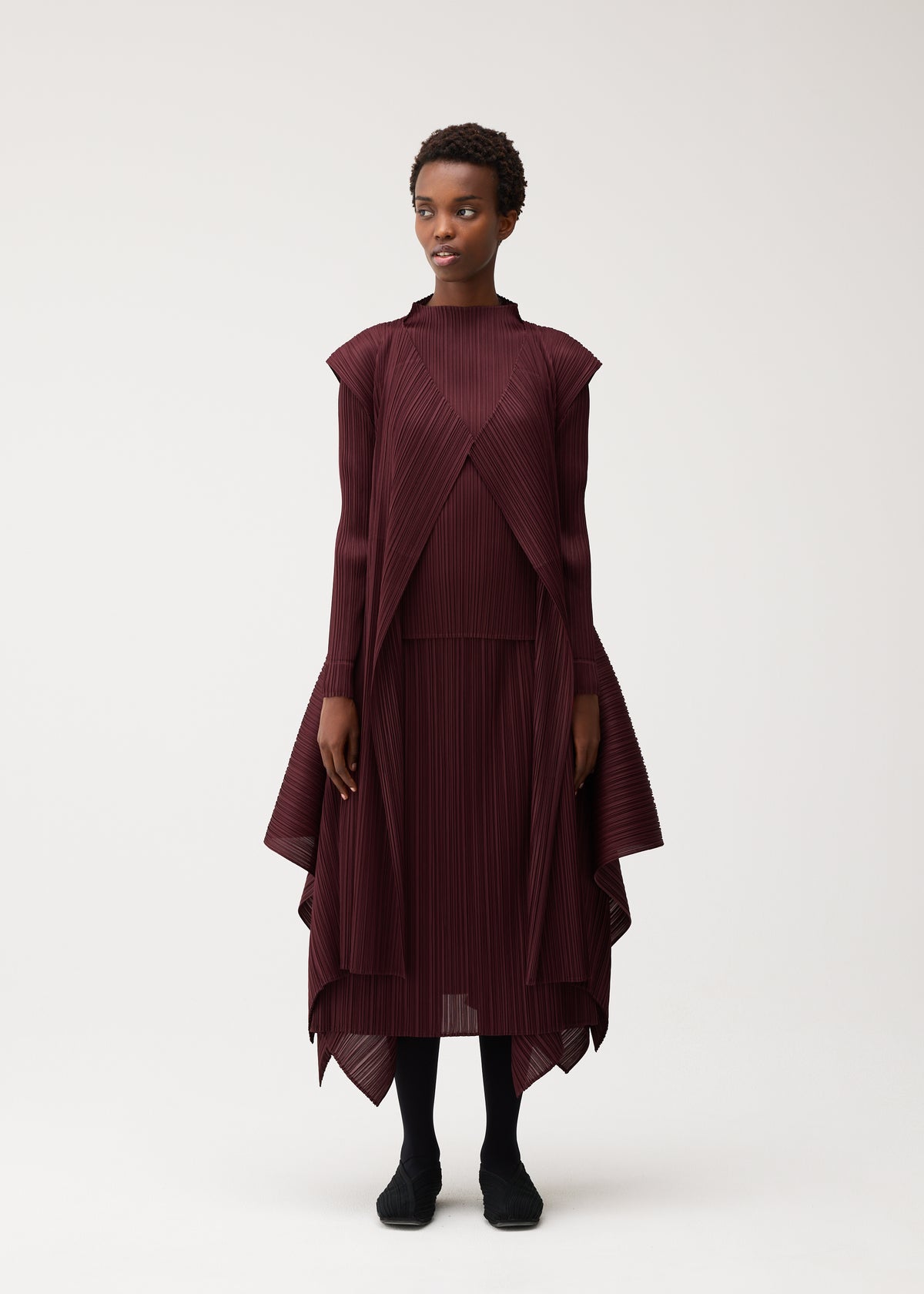 Zoomed view A woman with short hair poses against a plain white background wearing the FLUTTER dress by PLEATS PLEASE ISSEY MIYAKE—a burgundy, pleated, asymmetrical design with long sleeves and distinctive fabric details. She completes the look with black shoes.