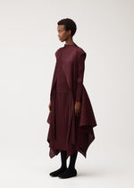 A person stands sideways wearing the FLUTTER dress by PLEATS PLEASE ISSEY MIYAKE, a burgundy, pleated, asymmetrical piece with long sleeves and a draped vest featuring a unique fabric design. They have short curly hair, black leggings, and black shoes, set against a plain light background.