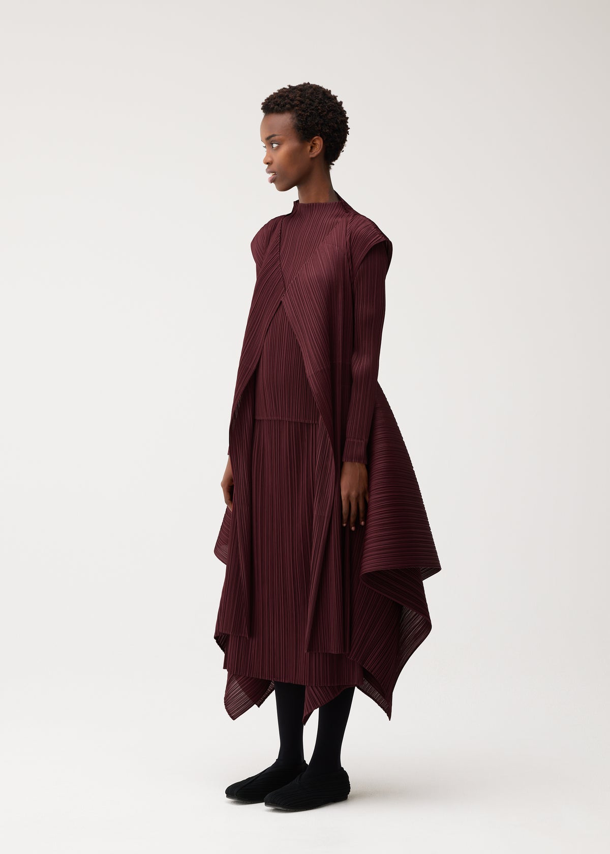 Zoomed view A person stands sideways wearing the FLUTTER dress by PLEATS PLEASE ISSEY MIYAKE, a burgundy, pleated, asymmetrical piece with long sleeves and a draped vest featuring a unique fabric design. They have short curly hair, black leggings, and black shoes, set against a plain light background.