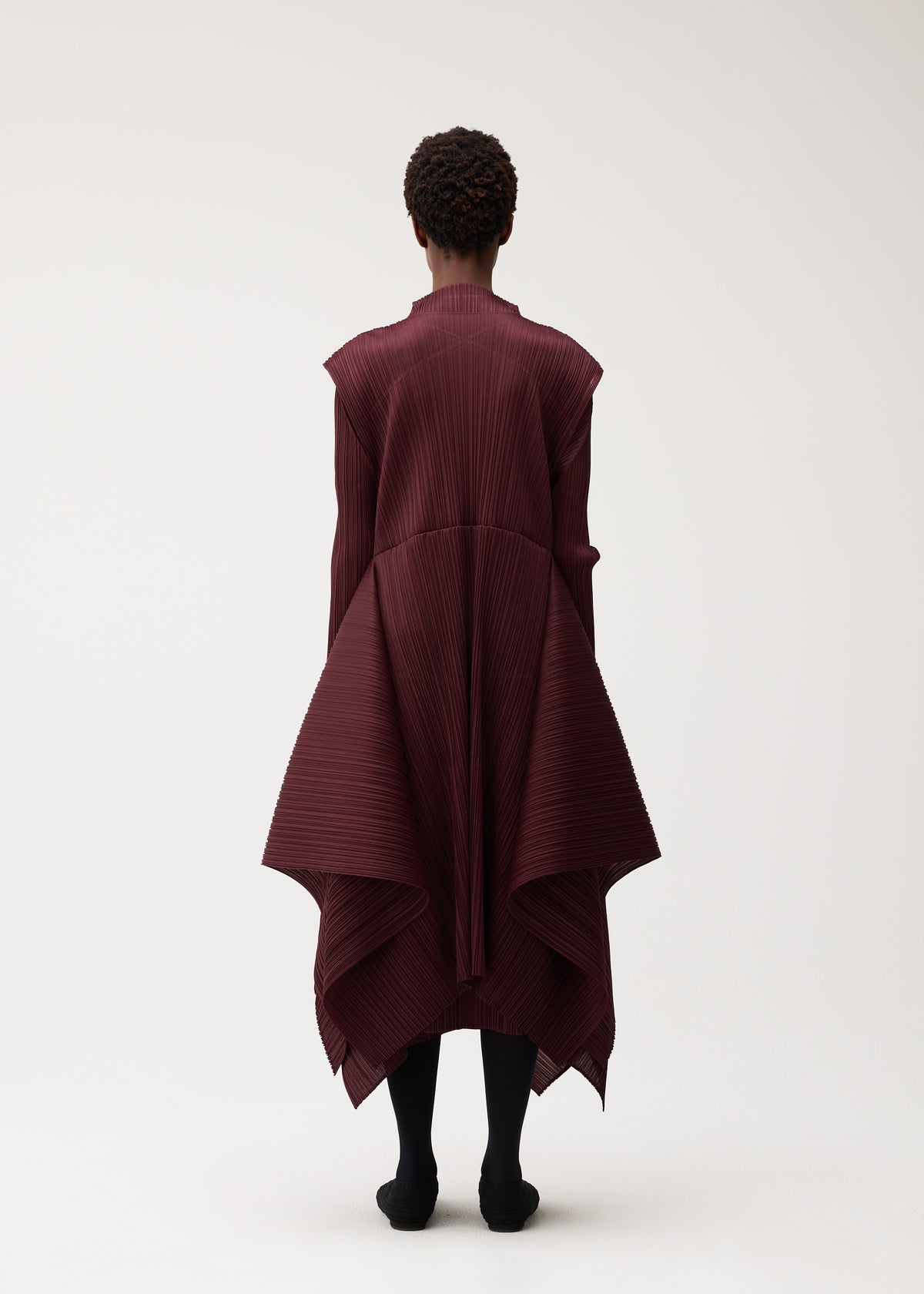 A person stands against a plain light background, seen from behind, wearing the PLEATS PLEASE ISSEY MIYAKE FLUTTER dress—a burgundy pleated asymmetrical design featuring draped panels and a distinctive fabric pattern. Their arms rest relaxed at their sides, paired with black tights. view 8 of 9