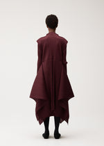 A person stands against a plain light background, seen from behind, wearing the PLEATS PLEASE ISSEY MIYAKE FLUTTER dress—a burgundy pleated asymmetrical design featuring draped panels and a distinctive fabric pattern. Their arms rest relaxed at their sides, paired with black tights.