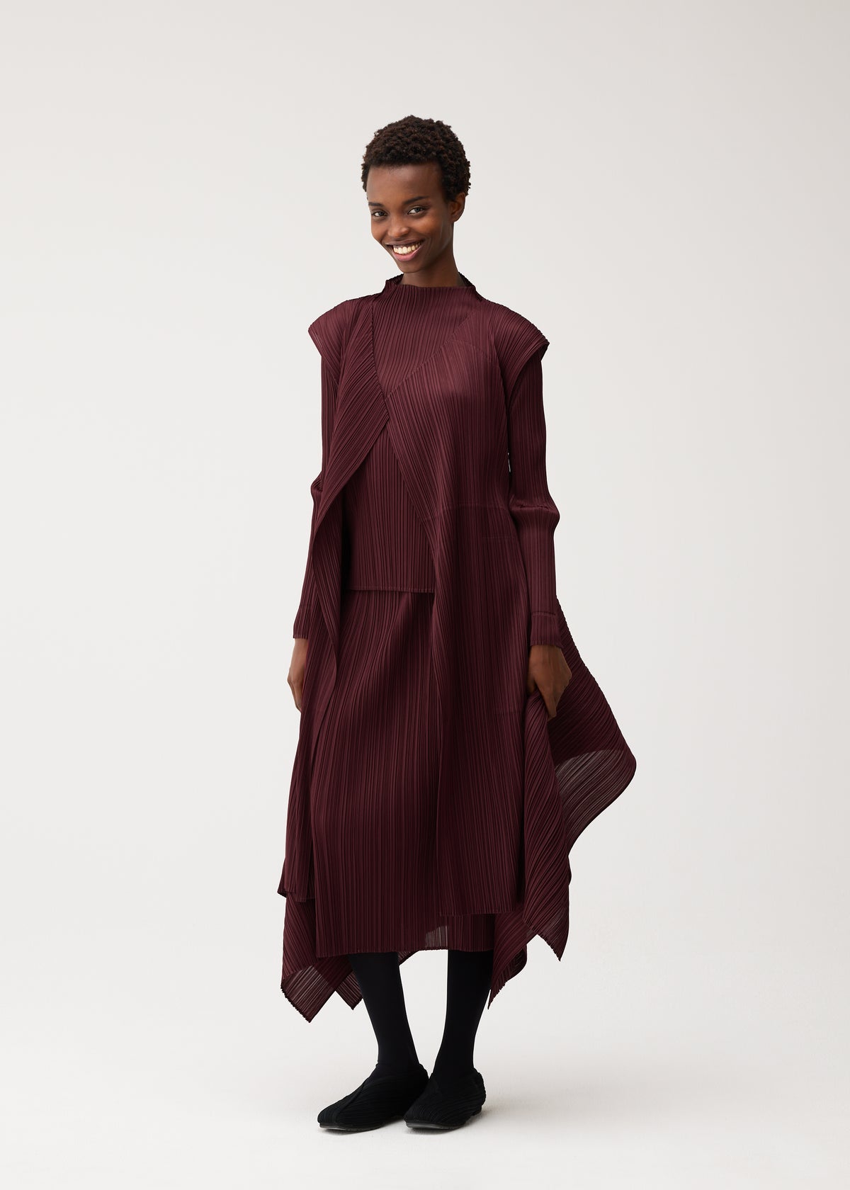 Zoomed view A woman stands smiling against a plain light background, wearing the FLUTTER long burgundy pleated dress with asymmetrical layers and a draped vest featuring a unique fabric design from PLEATS PLEASE ISSEY MIYAKE, paired with black tights and black shoes.