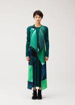 A person stands facing forward in the ORBIT pleated dress by PLEATS PLEASE ISSEY MIYAKE, featuring sheer overlapping green, blue, and black shapes, paired with black boots against a plain light background.