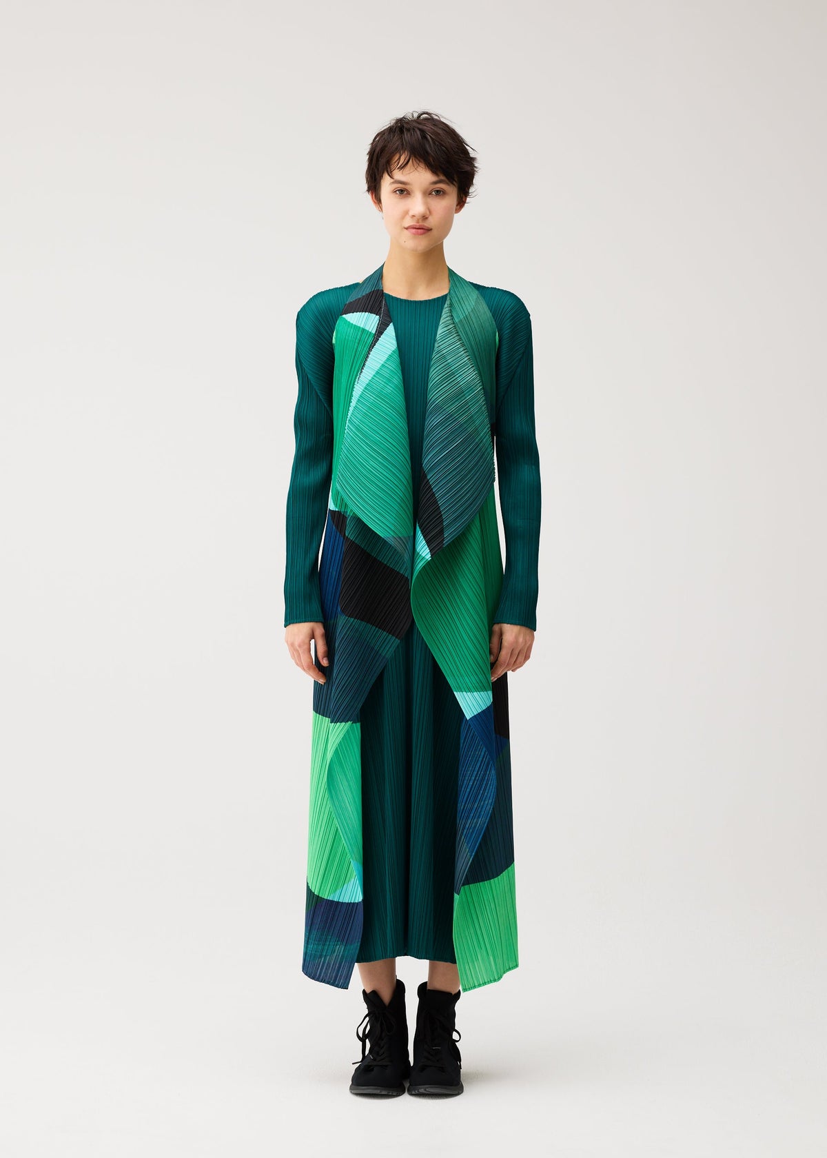 Zoomed view A person stands facing forward in the ORBIT pleated dress by PLEATS PLEASE ISSEY MIYAKE, featuring sheer overlapping green, blue, and black shapes, paired with black boots against a plain light background.