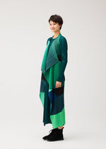 A person stands sideways, smiling, wearing the ORBIT long vest by PLEATS PLEASE ISSEY MIYAKE with green and blue overlapping shapes and black shoes against a plain light background.