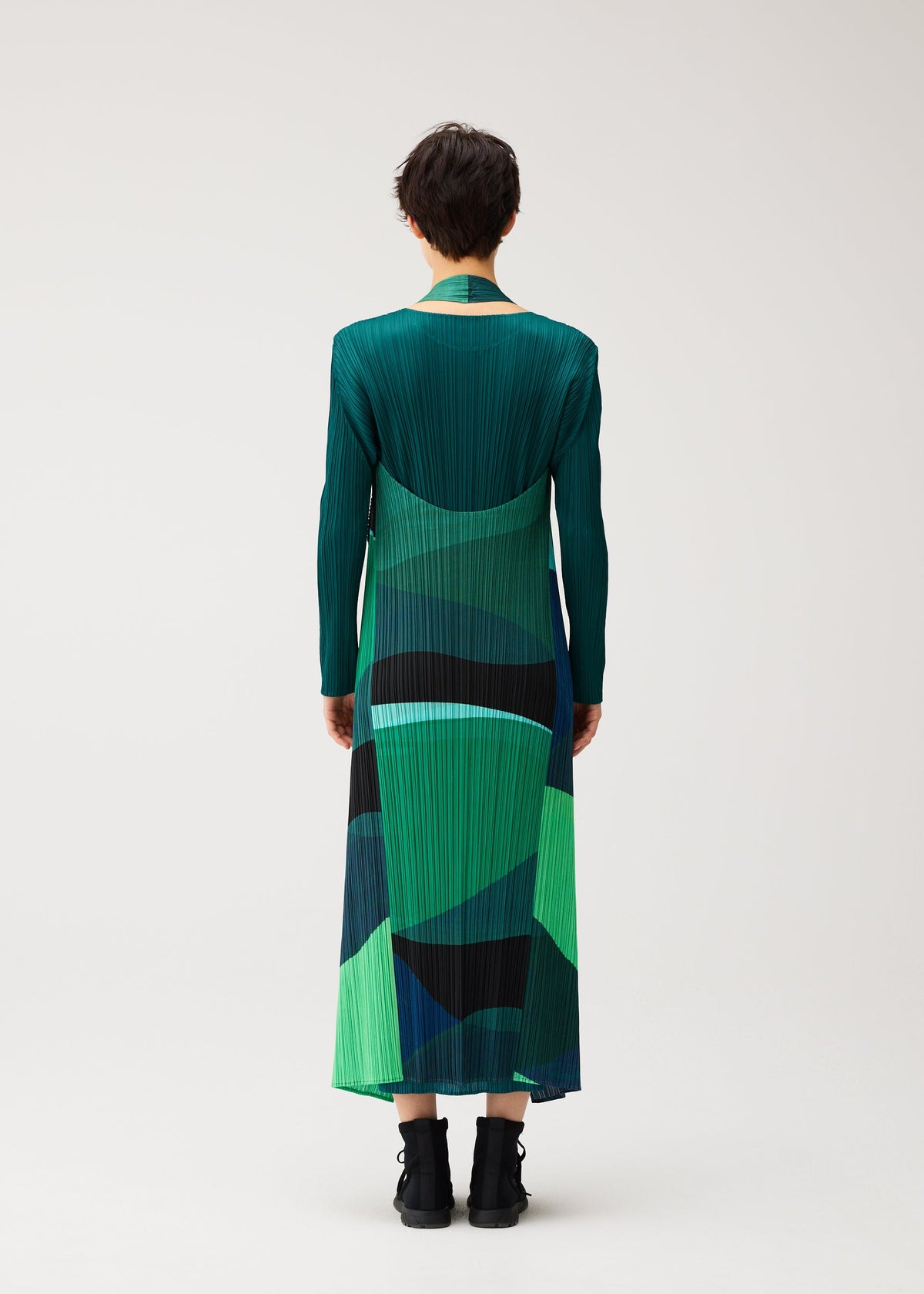 Zoomed view A person stands with their back to the camera, wearing the ORBIT long-sleeved pleated dress by PLEATS PLEASE ISSEY MIYAKE in sheer green and blue, paired with black ankle boots against a plain light background.