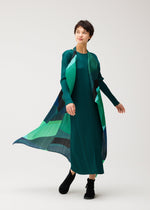 A person stands against a plain light background, wearing the ORBIT dress by PLEATS PLEASE ISSEY MIYAKE—its flowing sheer green panels with blue and black geometric patterns billow as they move, paired with black shoes.