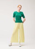 A woman with red hair and light skin stands against a plain white background, hands behind her back, slightly smiling, wearing the PLEATS PLEASE ISSEY MIYAKE MONTHLY COLORS : JULY ribbed green top and colorful wide yellow pants with pockets.