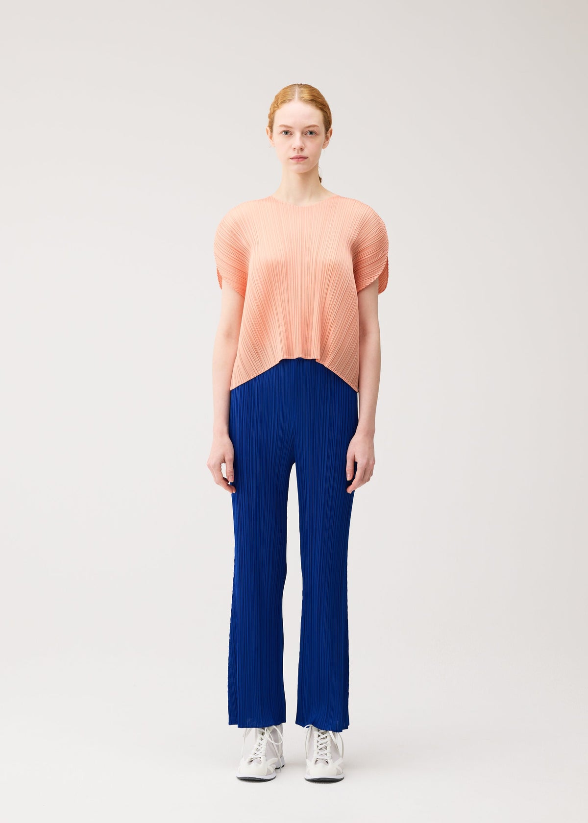 A woman with light skin and her hair pulled back wears the MONTHLY COLORS : JULY pleated top and colorful flare pants by PLEATS PLEASE ISSEY MIYAKE, paired with white sneakers, standing against a plain background. view 7 of 10