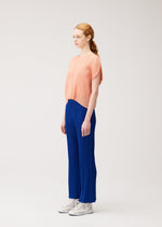 A woman with red hair in a ponytail models the PLEATS PLEASE ISSEY MIYAKE MONTHLY COLORS : JULY collection—she wears a loose peach pleated top and bright blue flare pants with pockets, styled with white shoes against a plain light background.