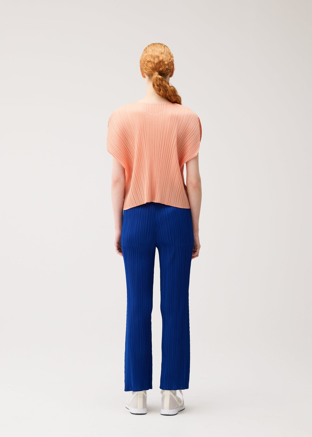 A person with red hair in a low ponytail faces away, wearing MONTHLY COLORS : JULY by PLEATS PLEASE ISSEY MIYAKE—featuring a loose, pleated peach top and colorful flare pants with pockets—against a plain light background. view 9 of 10