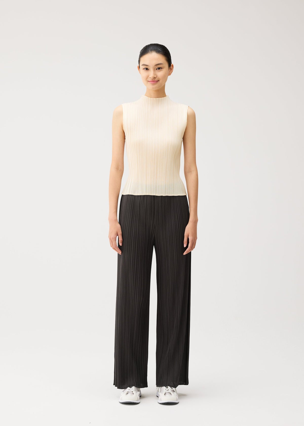A woman stands against a plain white background, dressed in PLEATS PLEASE ISSEY MIYAKE’s MONTHLY COLORS : AUGUST collection. She wears a sleeveless, light beige top paired with loose black straight pants and white sneakers. Smiling softly with her hair tied back, she effortlessly exemplifies understated summer style. view 8 of 11