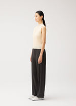 A woman with long dark hair in a ponytail stands sideways against a plain white background, wearing the effortlessly chic MONTHLY COLORS : AUGUST wide black pleated pants from PLEATS PLEASE ISSEY MIYAKE, paired with a sleeveless beige top and white shoes—an ideal summer outfit.