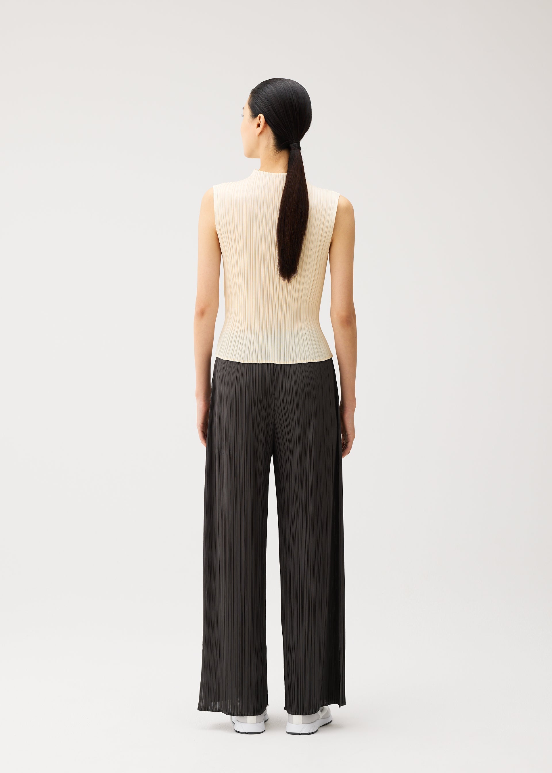 Zoomed view A person stands facing away, wearing the MONTHLY COLORS : AUGUST sleeveless, cream-colored top and loose, black pleated straight pants from PLEATS PLEASE ISSEY MIYAKE, paired with white shoes. Their straight, dark hair is tied in a low ponytail. Perfect for summer wear, this ensemble by PLEATS PLEASE ISSEY MIYAKE features striking colors that stand out against the plain, light background.