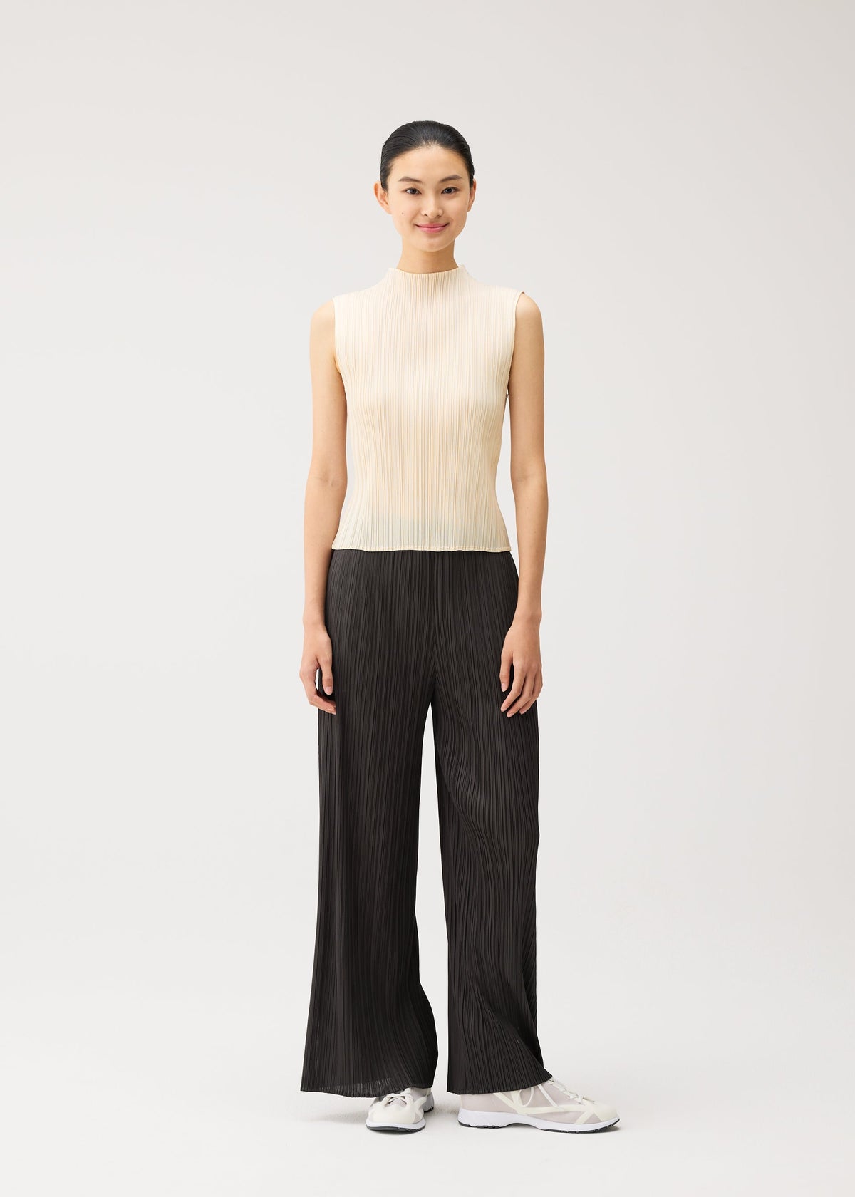 A woman stands against a plain background, smiling with her hair tied back, wearing the MONTHLY COLORS : AUGUST sleeveless beige top and wide-leg black pleated pants from PLEATS PLEASE ISSEY MIYAKE—an effortlessly stylish summer ensemble—paired with crisp white sneakers. view 11 of 11