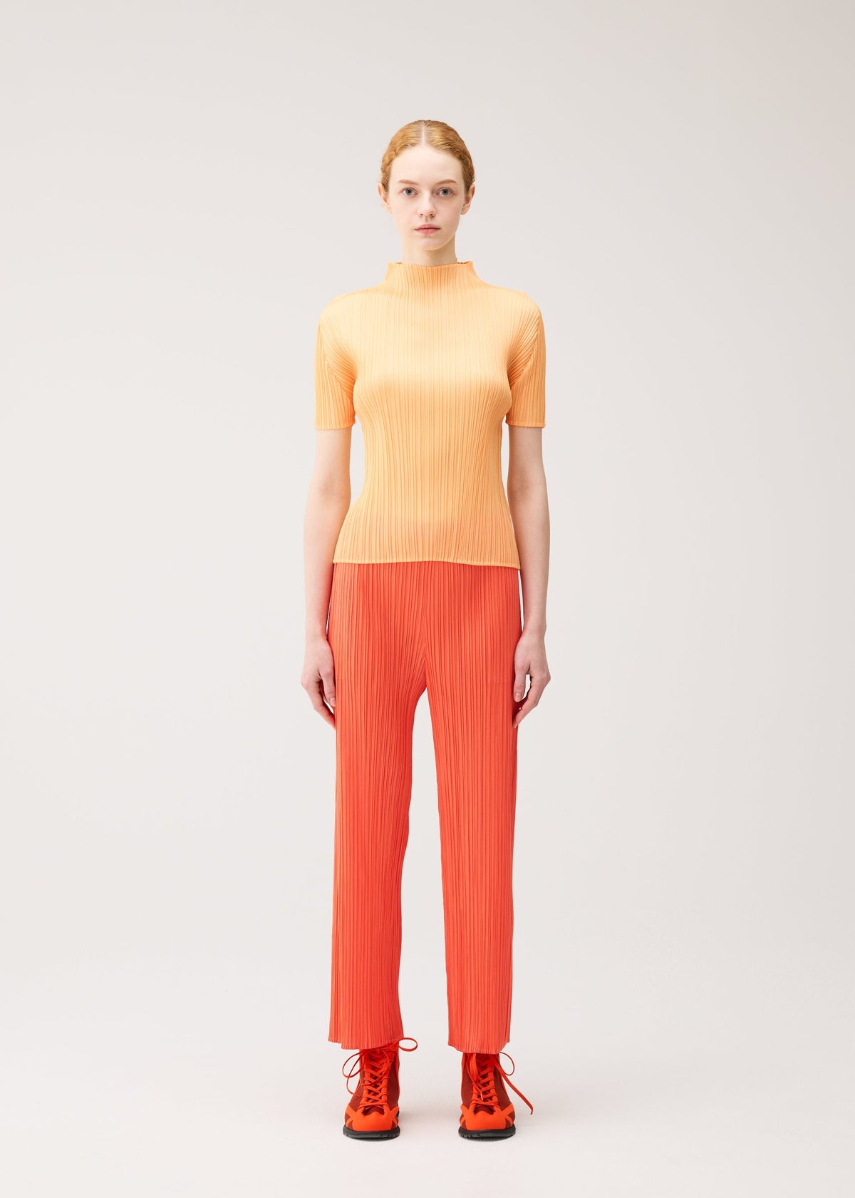 A woman with light skin and red hair in a ponytail models the MONTHLY COLORS : SEPTEMBER collection by PLEATS PLEASE ISSEY MIYAKE, wearing a short-sleeved, ribbed orange top paired with red straight pants and lace-up boots. She stands against a plain light background, her ensemble’s color palette creating a striking yet harmonious effect. view 7 of 10